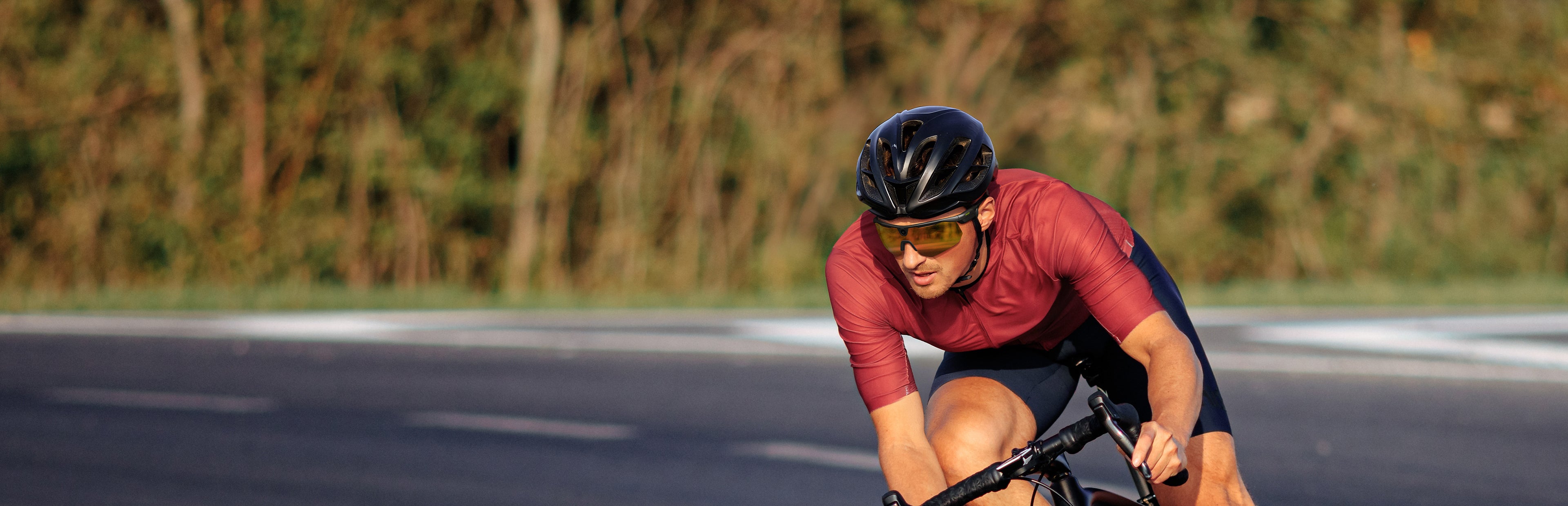 A cyclist riding on a paved road wearing a black aerodynamic cycling helmet and amber-tinted BLEEQUP Ranger AI Sports Camera Glasses (outdoor UGC scene, late afternoon, grassy background)